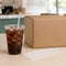 A clear plastic cup filled with iced cola sits on a light wood table next to a large brown cardboard box and several clear protective straw sleeves.