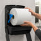 A person's hands are shown loading a large roll of white paper towels into a black wall-mounted dispenser with a blue pull tab, set against a light gray tiled bathroom wall.