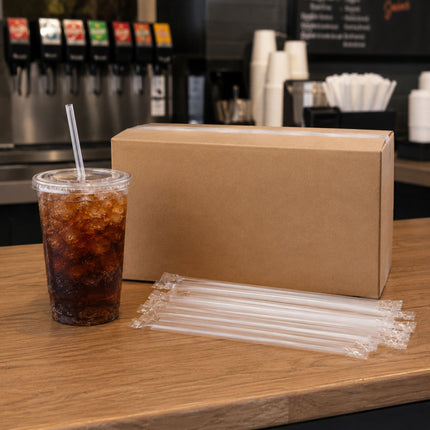 A clear plastic straw set displayed on a wooden counter next to a cardboard box and a iced cola cup with a clear straw, with beverage dispensers blurred in the background.