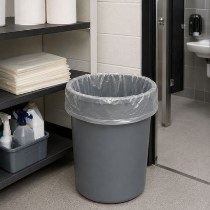 A gray plastic trash can with a clear plastic liner, placed on a tiled bathroom floor next to a shelf with cleaning supplies.