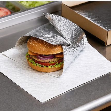A burger wrapped in a crinkled silver foil sleeve, resting on a white paper napkin on a stainless steel counter next to a cardboard box.