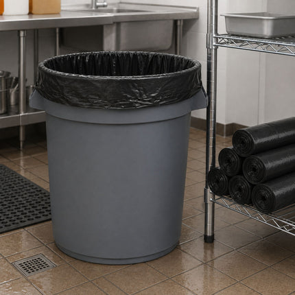 A large gray plastic trash can with a black plastic liner, placed on a tiled floor in a commercial kitchen or utility space next to a metal shelving unit.