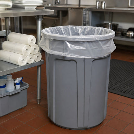 A large gray plastic trash can with a clear plastic liner, placed on a tiled kitchen floor next to a metal table holding rolls of liner paper.