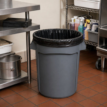 A gray plastic trash can with a black liner, placed on a tiled floor in a commercial kitchen setting next to stainless steel shelves.