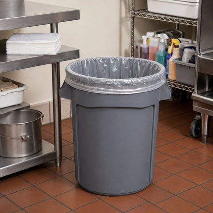 A gray plastic trash can with a clear plastic liner, placed on a brown tiled floor in a commercial kitchen setting with stainless steel shelves and equipment visible.
