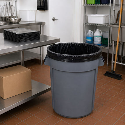 A gray plastic trash can with a black liner, placed on a tiled kitchen or utility floor next to stainless steel worktables and shelving.