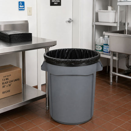 A gray plastic trash can with a black liner, placed on a tiled floor in a commercial kitchen setting.