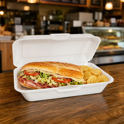 A large sub sandwich in a white foam take-out container with potato chips, sitting on a wooden table in a blurred restaurant or cafe setting.